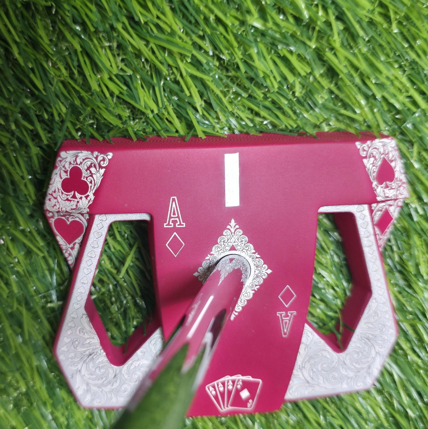 Detailed front view of a custom milled, deep matte red mallet putter head. Intricate playing card suit (Hearts, Diamonds, Spades, Clubs) engravings in satin silver. Features a fanned card graphic, "Ace of Diamonds" symbols, central hole, and center alignment aid. Engraved hearts on corners. ngled top-down view of a custom red mallet putter head resting on green artificial turf, showing the detailed silver scrollwork wings with small suit symbols.
