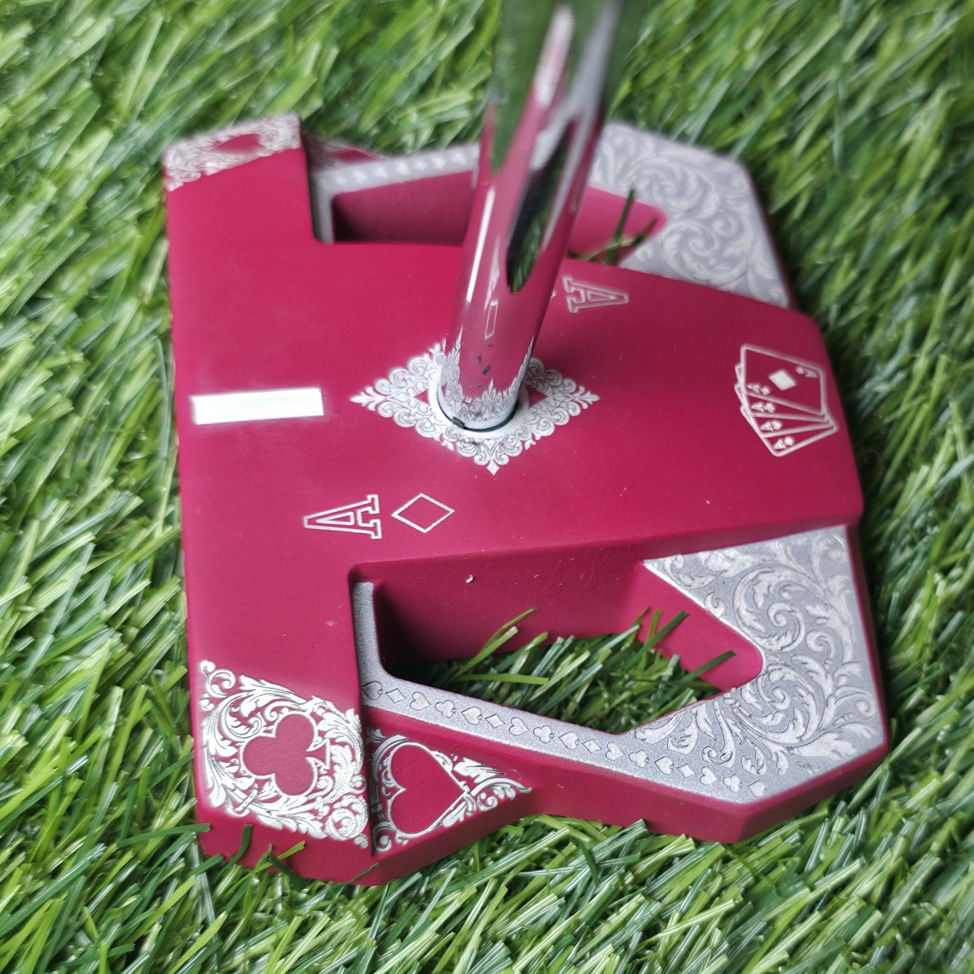 Detailed front view of a custom milled, deep matte red mallet putter head. Intricate playing card suit (Hearts, Diamonds, Spades, Clubs) engravings in satin silver. Features a fanned card graphic, "Ace of Diamonds" symbols, central hole, and center alignment aid. Engraved hearts on corners. ngled top-down view of a custom red mallet putter head resting on green artificial turf, showing the detailed silver scrollwork wings with small suit symbols.