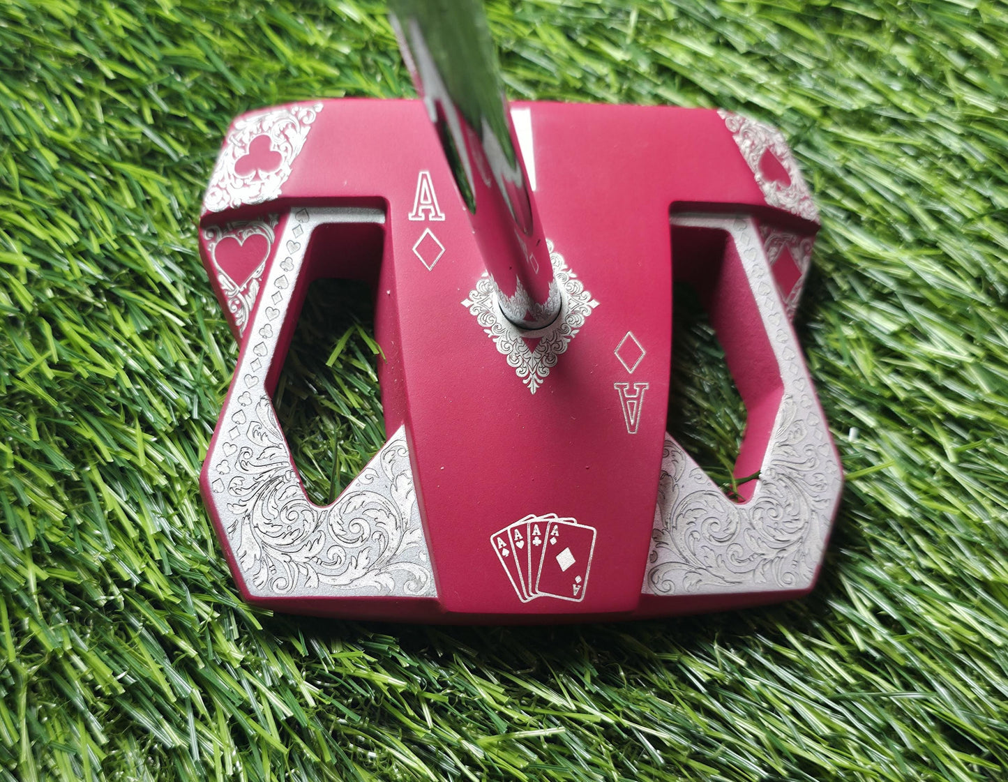 Detailed front view of a custom milled, deep matte red mallet putter head. Intricate playing card suit (Hearts, Diamonds, Spades, Clubs) engravings in satin silver. Features a fanned card graphic, "Ace of Diamonds" symbols, central hole, and center alignment aid. Engraved hearts on corners. ngled top-down view of a custom red mallet putter head resting on green artificial turf, showing the detailed silver scrollwork wings with small suit symbols.