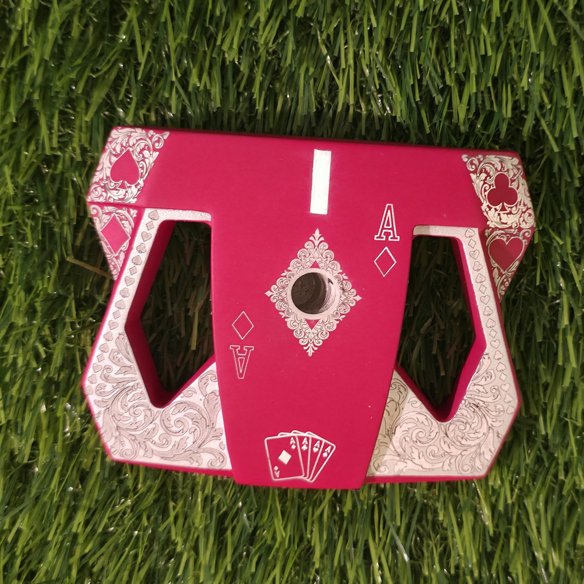 Detailed front view of a custom milled, deep matte red mallet putter head. Intricate playing card suit (Hearts, Diamonds, Spades, Clubs) engravings in satin silver. Features a fanned card graphic, "Ace of Diamonds" symbols, central hole, and center alignment aid. Engraved hearts on corners. ngled top-down view of a custom red mallet putter head resting on green artificial turf, showing the detailed silver scrollwork wings with small suit symbols.