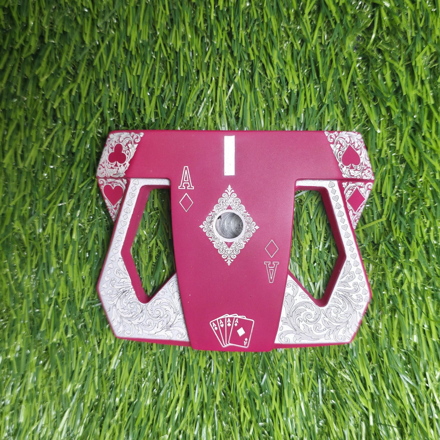 Detailed front view of a custom milled, deep matte red mallet putter head. Intricate playing card suit (Hearts, Diamonds, Spades, Clubs) engravings in satin silver. Features a fanned card graphic, "Ace of Diamonds" symbols, central hole, and center alignment aid. Engraved hearts on corners. ngled top-down view of a custom red mallet putter head resting on green artificial turf, showing the detailed silver scrollwork wings with small suit symbols.