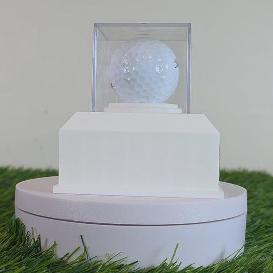 This custom golf ball display case rests on artificial green turf. It features a geometric 3D-printed base in matte black or crisp white. A clear acrylic cube sits on top to protect a standard white golf ball. On the front of the base is a rectangular brass metal plaque fastened with two silver screws. The plaque can be left blank or laser-engraved with personalized text like a Hole in One achievement, player name, date, and golf course. This beautiful display is perfect for sports memorabilia!!