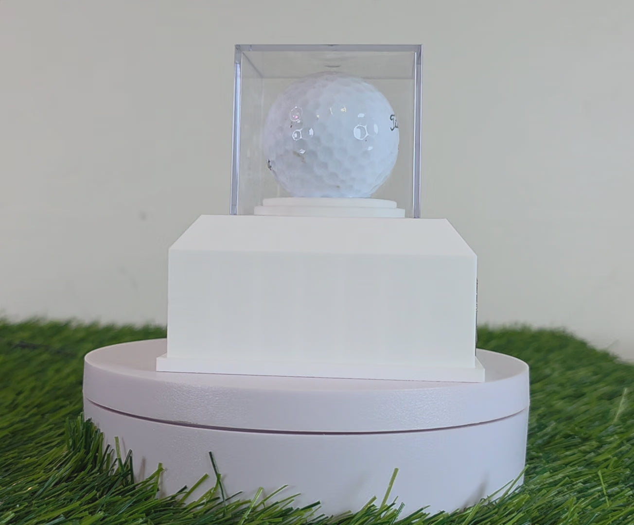 This custom golf ball display case rests on artificial green turf. It features a geometric 3D-printed base in matte black or crisp white. A clear acrylic cube sits on top to protect a standard white golf ball. On the front of the base is a rectangular brass metal plaque fastened with two silver screws. The plaque can be left blank or laser-engraved with personalized text like a Hole in One achievement, player name, date, and golf course. This beautiful display is perfect for sports memorabilia!!